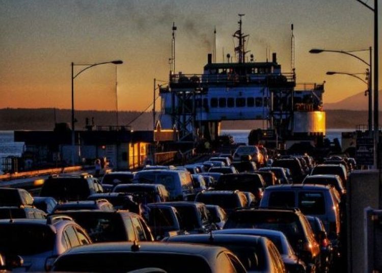 Vehicles lined up at a ferry terminal during sunset, with a silhouette of a large ferry in the background.