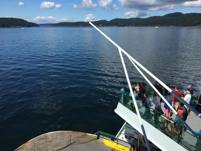 View from a ferry deck with people observing the water and distant forested coastline.
