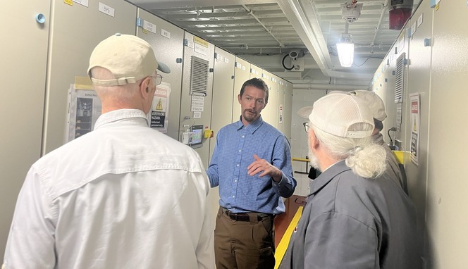 A man in a blue shirt explains something to three others in a corridor with electrical panels.