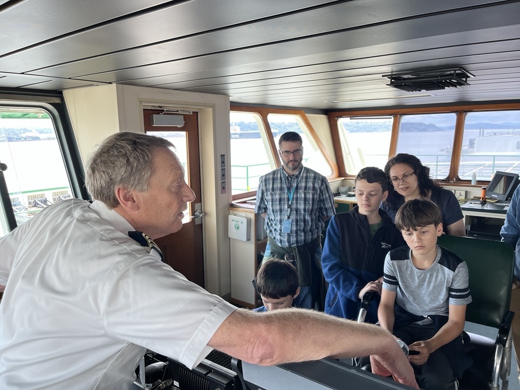 A man in a white uniform explains equipment to a group on a ship's bridge.