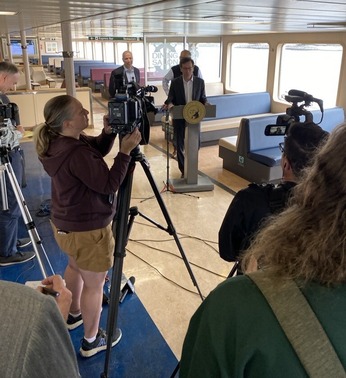 A press conference inside a ferry with a man speaking at a podium surrounded by journalists with cameras.