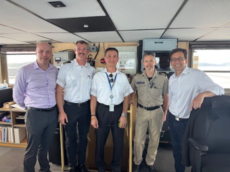 A group of five people standing in a command center room with control equipment behind them.