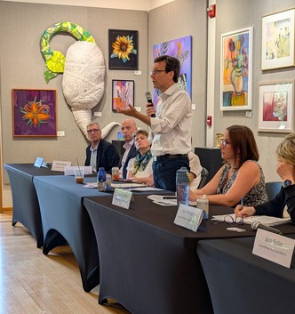 A person stands speaking at a panel discussion with artwork on the walls behind.