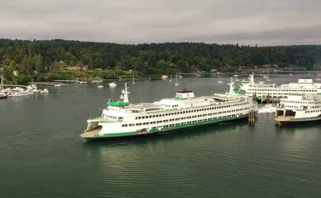 A white and green ferry boat sailing near a dock with forested shoreline in the background.