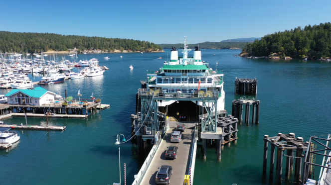 A ferry at a terminal with cars disembarking, surrounded by a marina with boats and forested shoreline.
