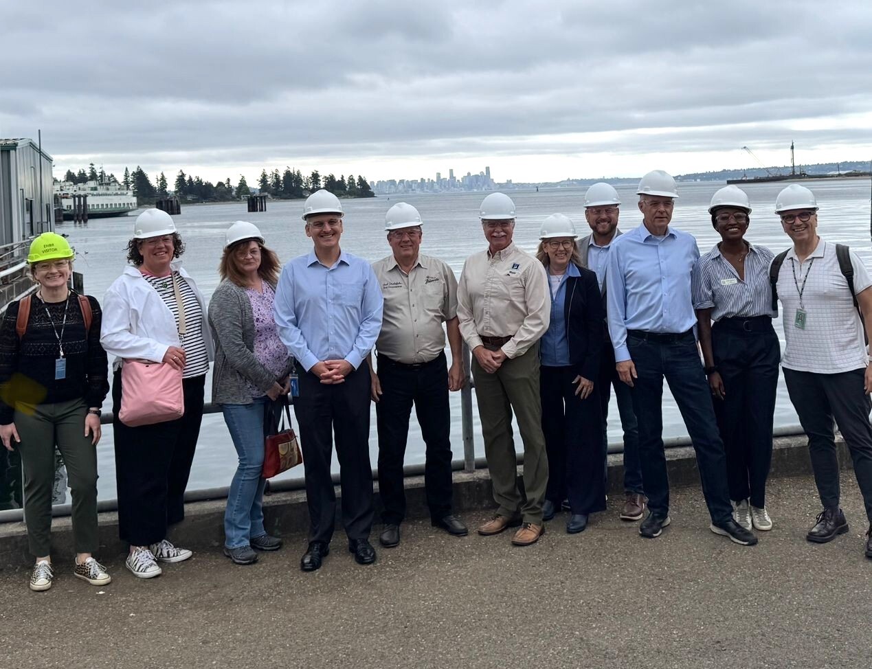 A group of 11 people wearing hard hats stand in front of a body of water with a city skyline in the background.