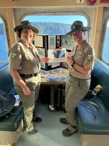 Two park rangers hold a map inside the passenger cabin of a ferry with brochures and a display board on a table behind them.