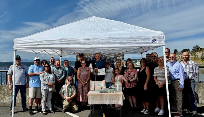 Group of people standing under a white tent by the ocean, with a table displaying items.