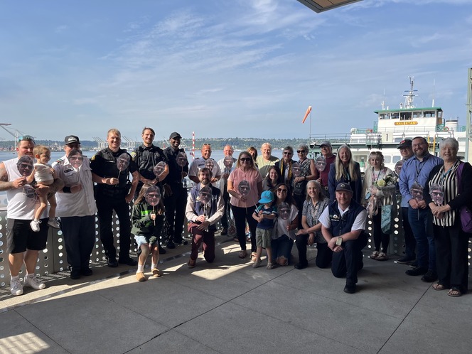 A group of people on a platform by the water, some holding face cutouts, with a ferry in the background on a sunny day.
