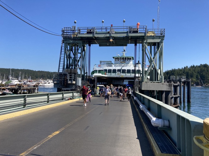 People walking off the ferry Tillikum docked at Friday Harbor on a sunny day, with a marina and forest visible in the background.