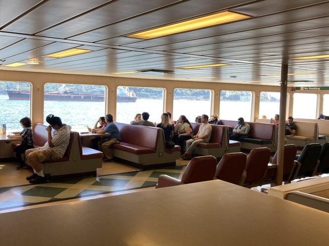 Passengers sitting in booths on a ferry with large windows showing a water view.
