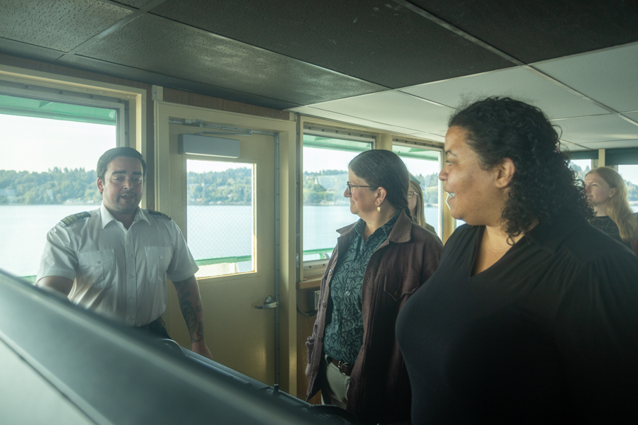 Three people in a ferry's wheelhouse with large windows overlooking water, engaged in conversation.