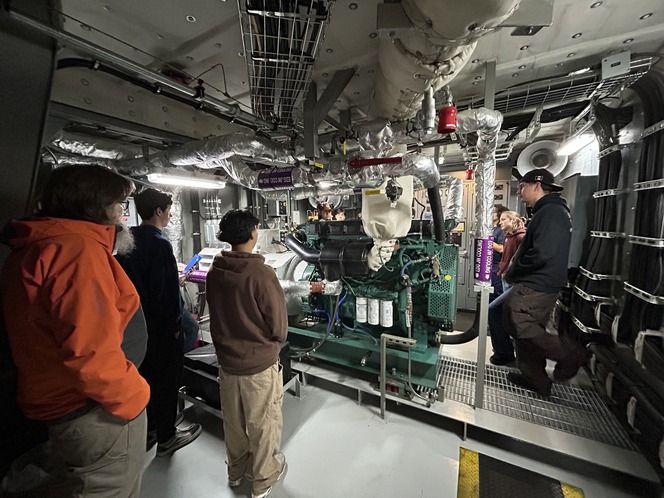 A group of people standing in a ferry's engine room with industrial machinery and equipment.