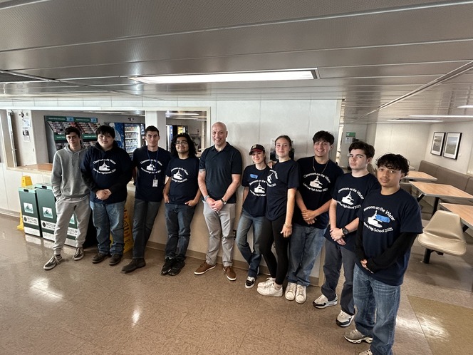 A group of ten people standing in a ferry's passenger cabin, most wearing navy blue t-shirts.