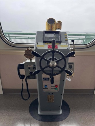 A ship's helm control with a steering wheel, electronic display, and brass compass, in front of a window showing the ocean.