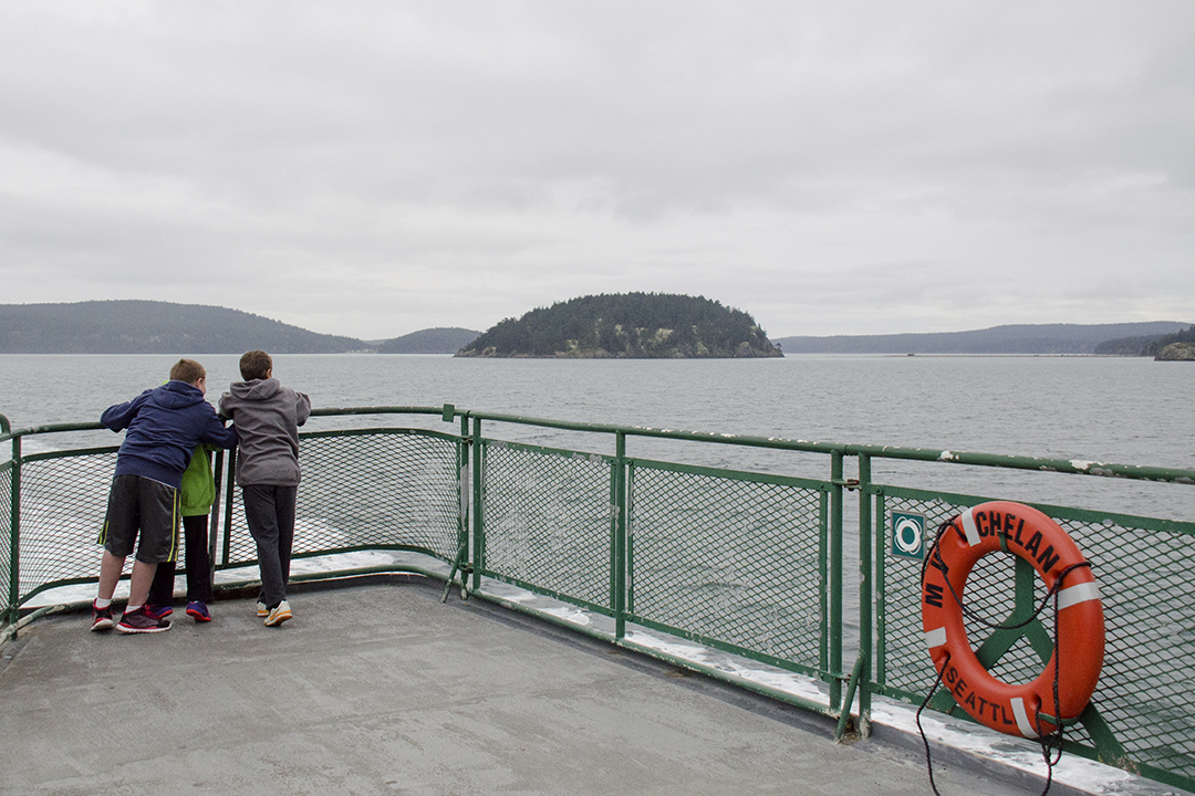 Children leaning on a ferry railing, looking at a distant island on an overcast day.