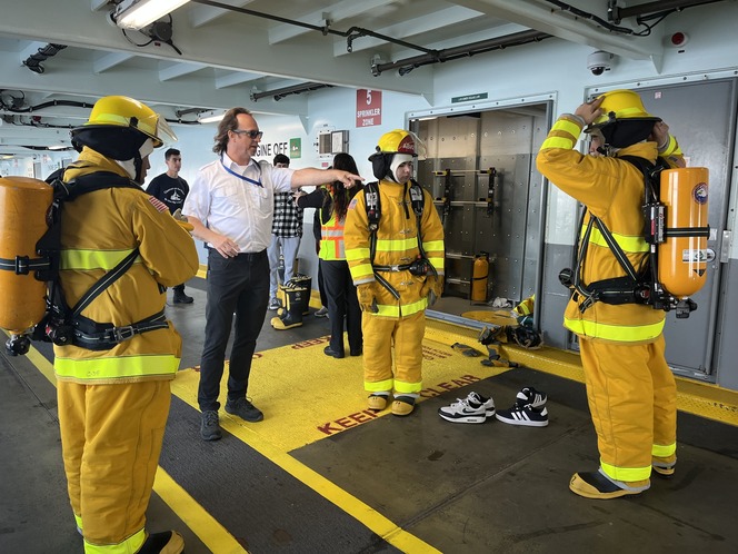 Three students in yellow firefighting gear listen to a person giving instructions on the car deck of a ferry.