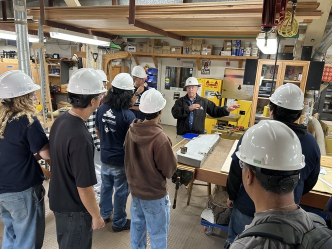 Individuals in hard hats listen to a demonstration in a woodworking workshop.