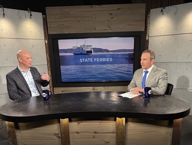 Two men in suits seated at a studio table, discussing in front of a large screen displaying "STATE FERRIES."