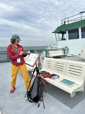 Person painting on a ferry deck with supplies on a bench and ferry's control house in the background.