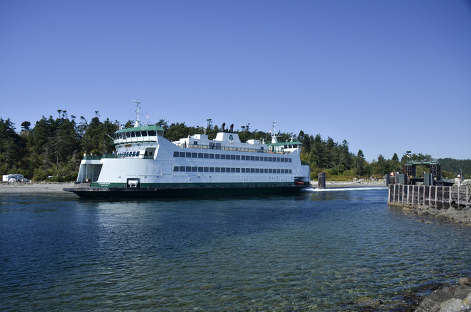 The ferry Kennewick travels through clear blue waters in Keystone Harbor with trees and Coupeville terminal in the background.