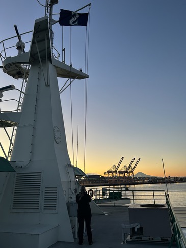 Ferry deck at sunset with a mast and Seattle Mariners flag, person silhouetted, T-Mobile Park, port cranes and distant mountain.