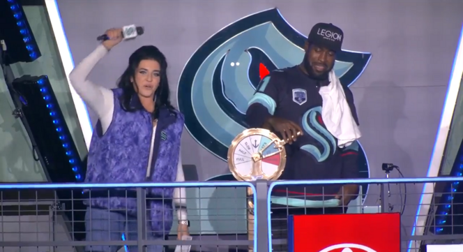 Two people on stage, one turning an engine telegraph and the other holding a microphone, with a Kraken logo in the background.