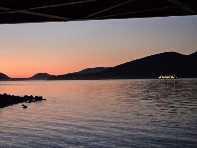 Sunset over calm water with silhouettes of hills and a lit ferry on the water.
