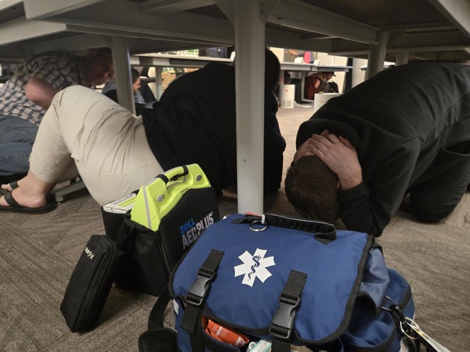 People crouching under tables, simulating an emergency drill. Emergency medical bags are in the foreground.