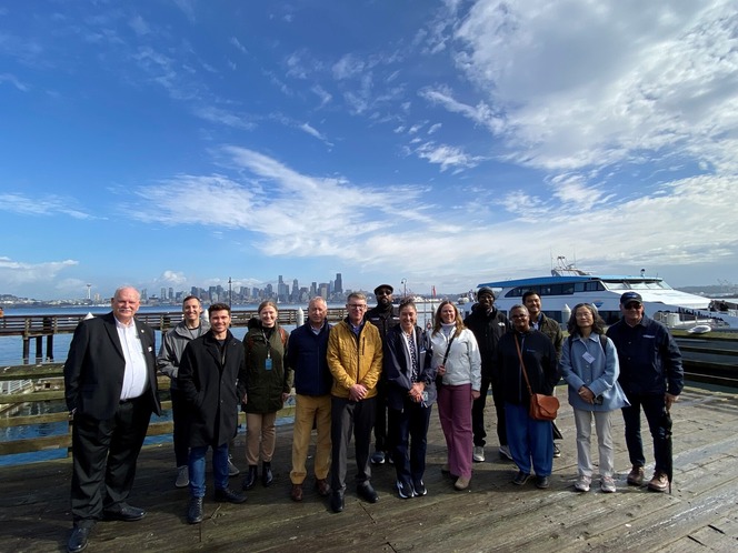 A group of 14 people stands on a dock with a city skyline and a ferry boat in the background under a partly cloudy sky.