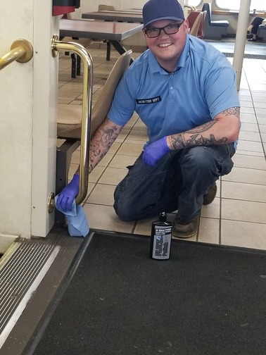 Person cleaning a brass handrail while crouching on a tiled floor, holding a blue cloth and wearing a blue polo, cap, and purple gloves.