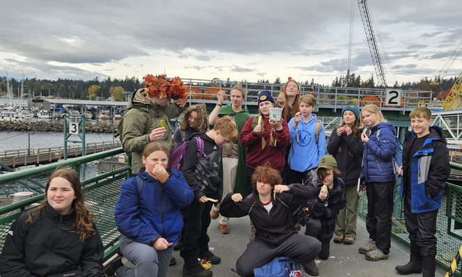 A group of teenagers on a ferry deck, with some holding pickles and a jar of olives, against an overcast marina backdrop.