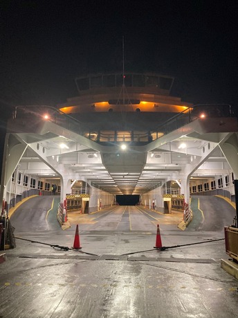 Rear night view of an illuminated ferry entrance with empty vehicle lanes and traffic cones.