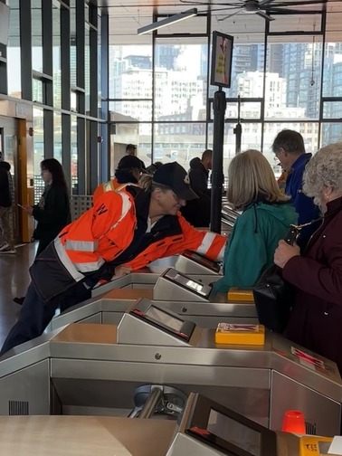 People at a transit station interacting with turnstiles, assisted by someone in a reflective orange jacket.