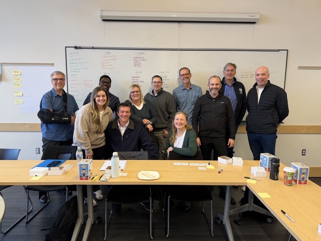 Group of people in a classroom, with a whiteboard and table filled with various items.