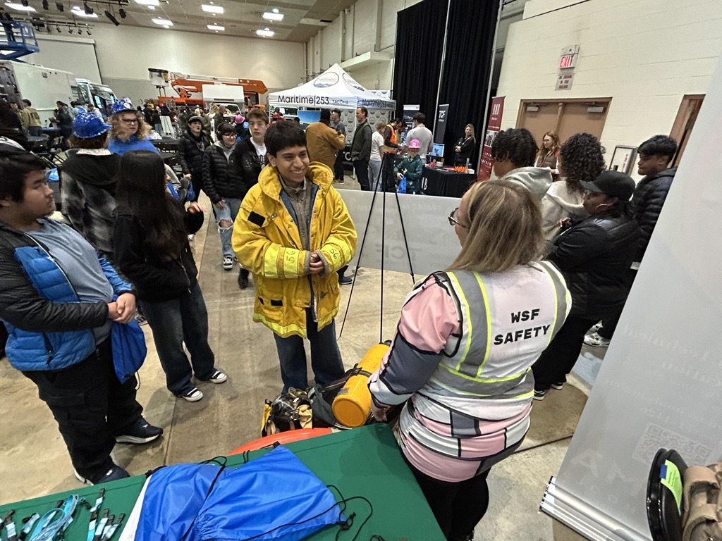People at an indoor event with booths and safety equipment. A person wears a yellow firefighter jacket, while another wears a "WSF SAFETY" vest.