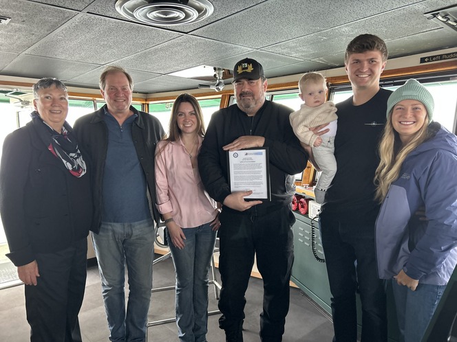 A group of seven people, including one holding a baby and another holding a framed document, pose in the wheelhouse of a ferry.