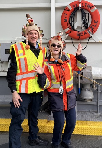 Two people in safety gear wearing turkey hats, standing in front of a life ring labeled "MV SEALTH SEATTLE."