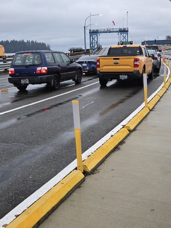 Vehicles lined up on a wet road near a ferry terminal, with yellow posts and curb separating the roadway from a sidewalk.