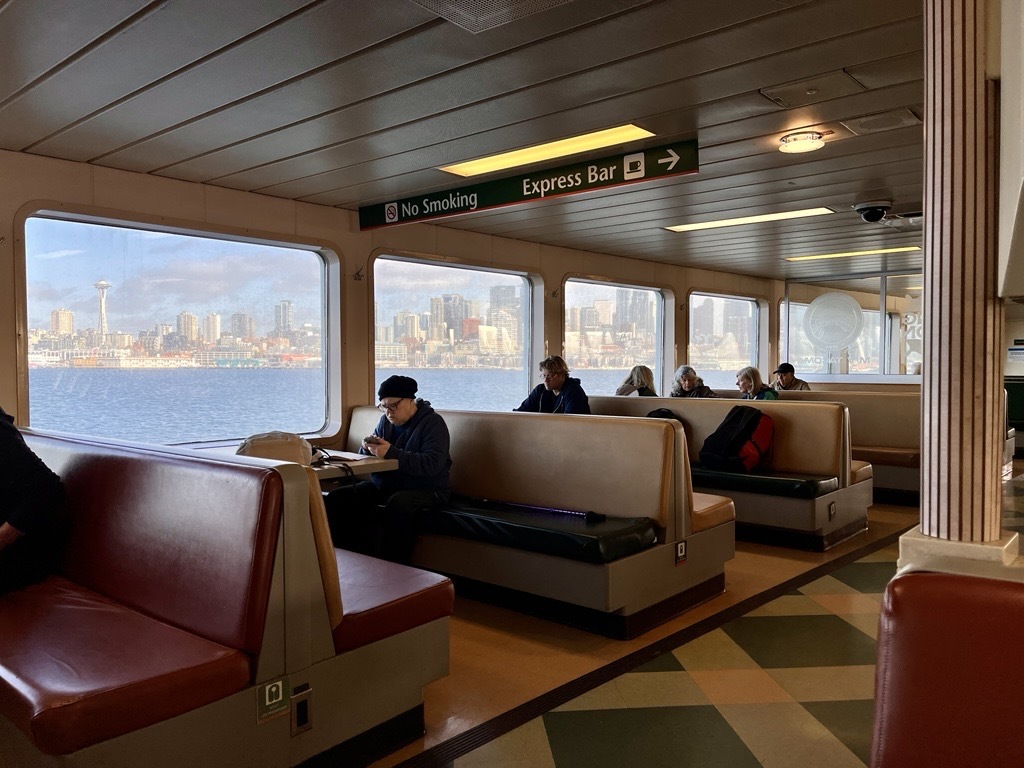 Interior of a ferry with passengers seated by windows showing a city skyline.