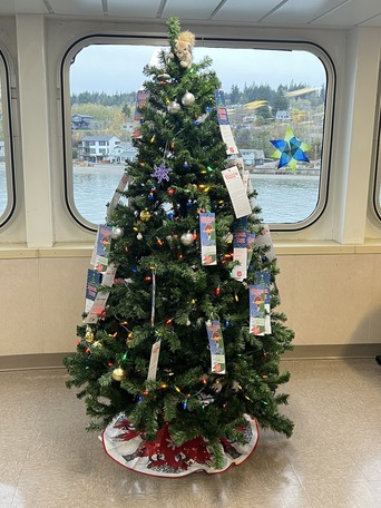 A decorated holiday tree by a window on a ferry with multicolored lights, informational tags and a stuffed toy squirrel up top.