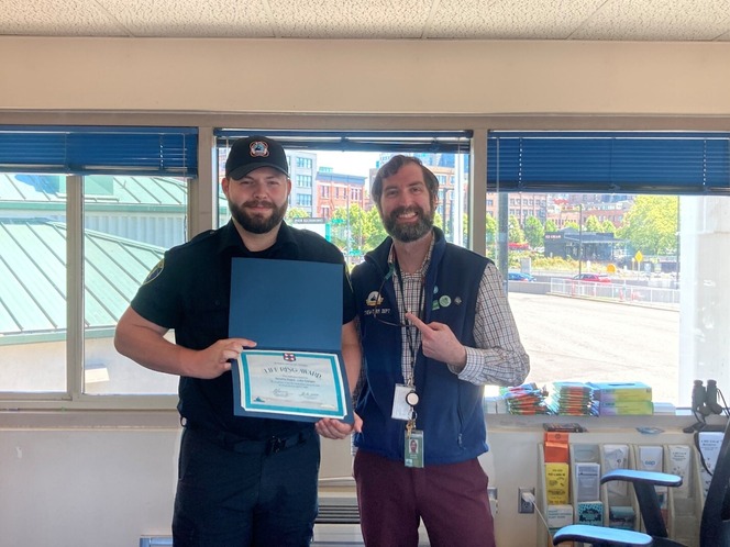 Two men stand in an office, one holding a certificate. Both are smiling.