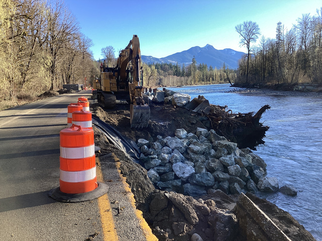 Crews work to rebuild an embankment along State Route 530 next to the Sauk River on a sunny day.