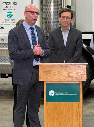 Two men in suits stand by a wooden lectern indoors, with a Washington Department of Transportation vehicle in the background.