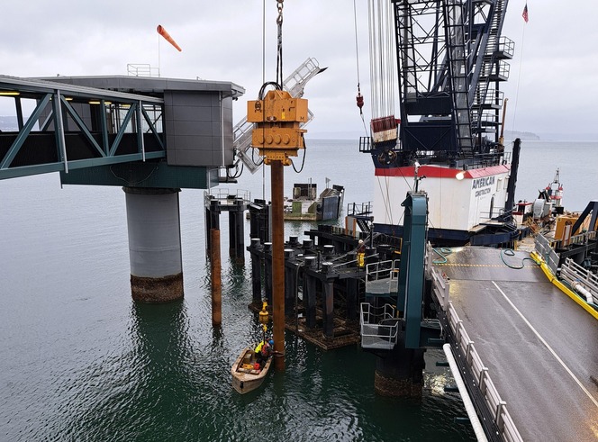 A maritime construction site with a large suspended drilling machine, a docked vessel with a lattice tower, and an over-water walkway.