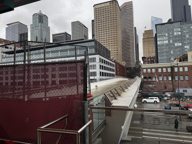A red mesh fence blocks off a pedestrian bridge, with a cityscape with tall buildings in the background and an overcast sky.