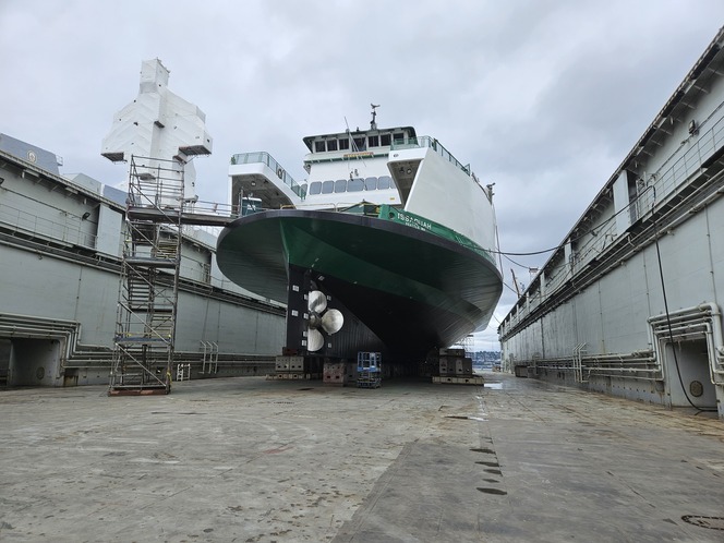 A large ferry in a dry dock, elevated on blocks with visible propellers, next to scaffolding under a cloudy sky.