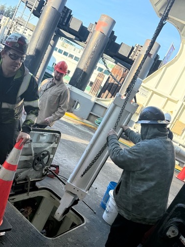 Workers on the car deck of a ferry with safety gear, managing equipment near an open hatch.