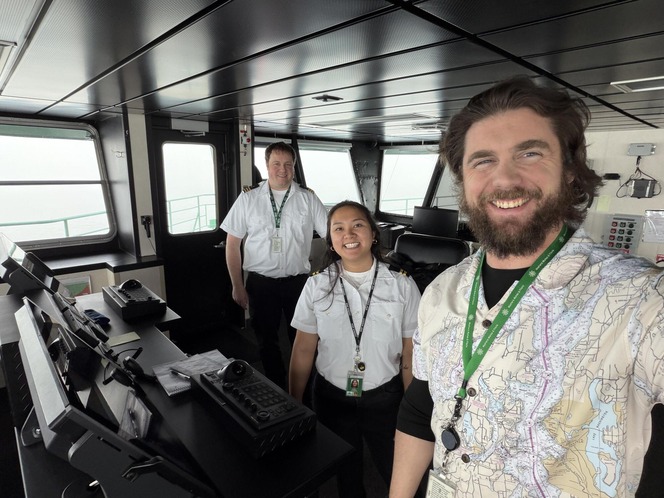 Three people smiling in a ferry's pilothouse, surrounded by control panels and windows.