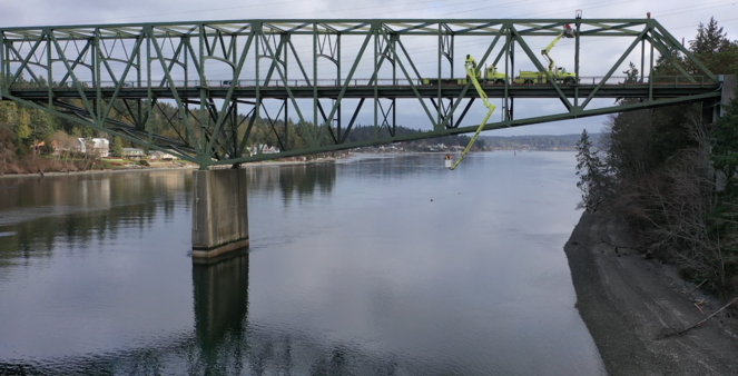A large green steel bridge spans a calm river, with a construction vehicle on it extending an arm down.